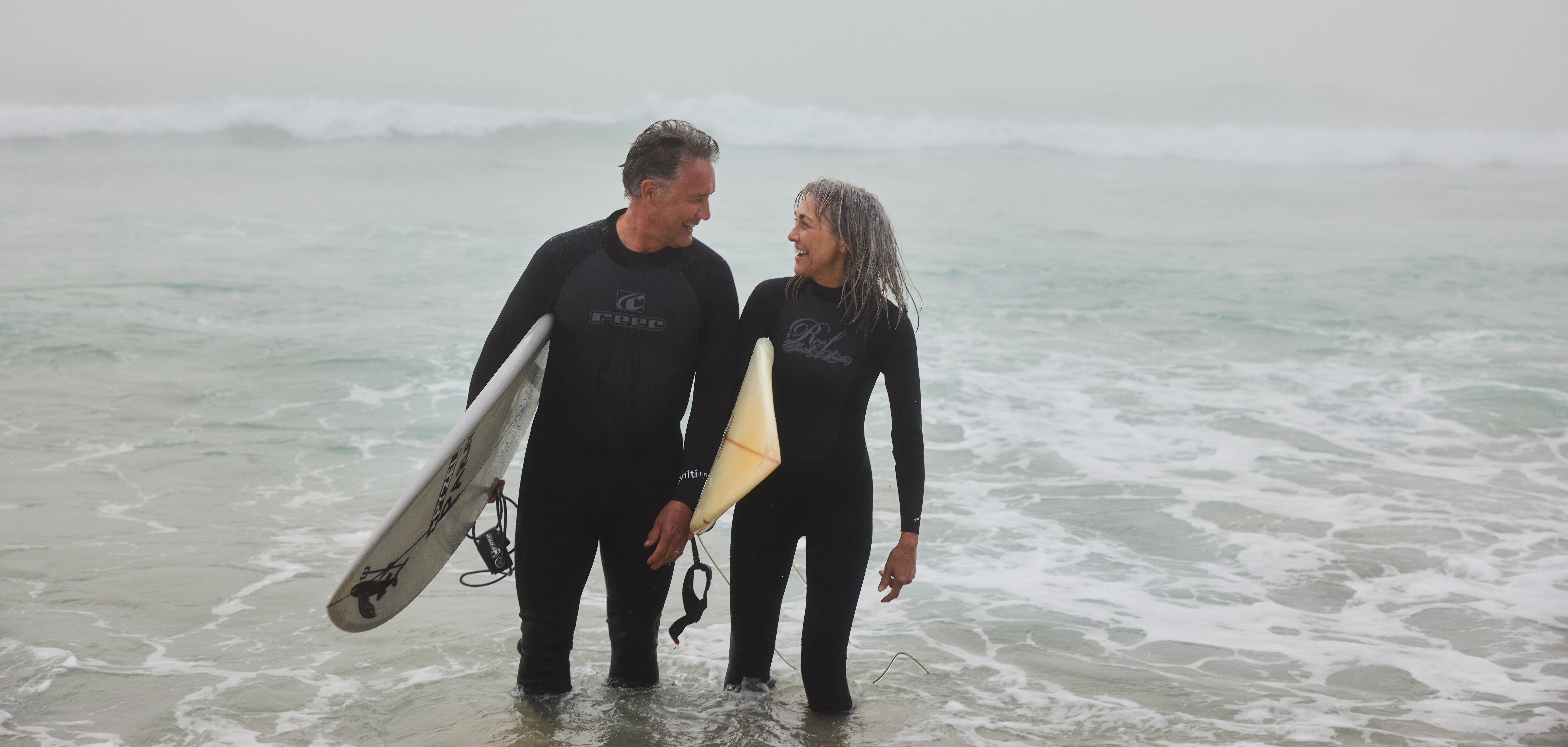 Older couple in wetsuits carrying a surfboard on the beach